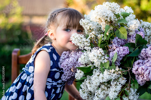 Little girl with blue eyes sniffing lilac flowers in a polka dot dress.
Horizontal closeup portrait of a little girl with a lilac.