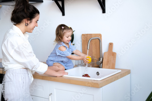 A beautiful young woman with her two year old daughter is washing fruit in the kitchen sink. Fresh bright fruits, apples, oranges, pears. Healthy food for children. The baby helps her mother.