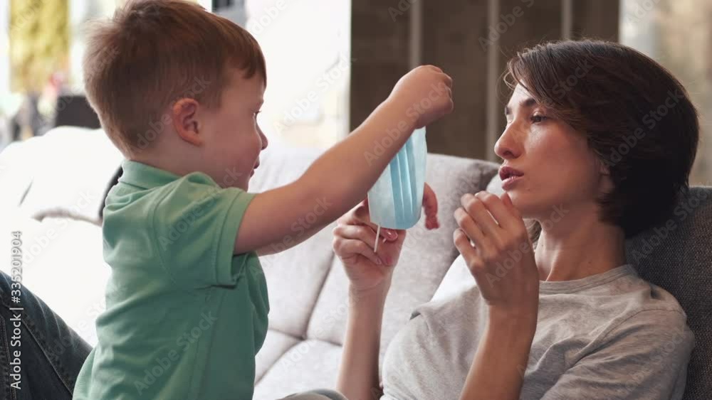 Pretty mother is playing with her son on a sofa at home. The woman tries to wear a mask. The parent is spending quarantine at home with children in self-isolation. Pretty mother is playing with her son on a sofa at home. The woman tries to wear a mask. The parent is spending quarantine at home with children in self-isolation.