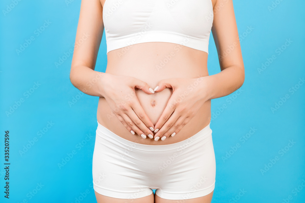 Close up of young pregnant woman making heart shape with her hands on the belly at blue background. Love to the future baby. Copy space