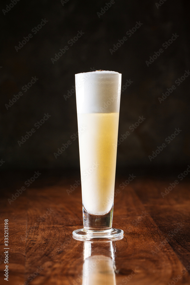 delicious light yellow sour foam cocktail in a long narrow glass and fresh lemon slice, dark wooden background, vertical, side view delicious light yellow sour foam cocktail in a long narrow glass and fresh lemon slice, dark wooden background, vertical, side view