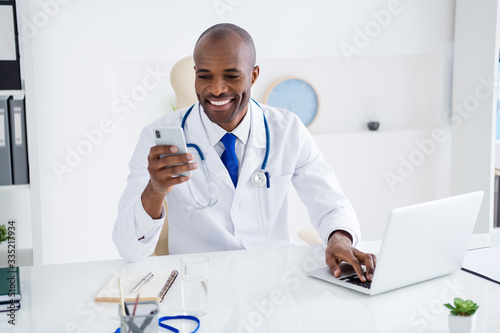 Photo of family doc dark skin guy watch notebook news write online consultation use telephone read patients email write prescription wear white lab coat sit chair office clinic indoors