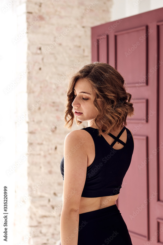 Fototapeta premium Young pretty woman with curly hair, wearing black shorts and top, is standing by burgundy wall. Morning fitness exercise in light sunny room
