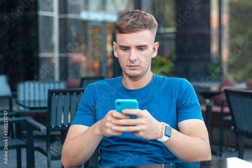 Serious handsome guy, young concentrated sad worried man looking at his cell mobile smart phone, typing message in terrace in cafe outdoors. Social media addiction. Gadgets, devices technology concept