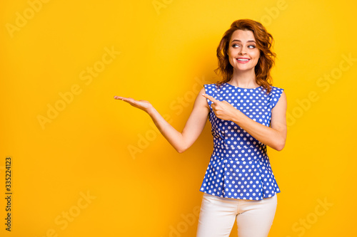Photo of attractive curly foxy business lady hold novelty product on open arm indicate finger presentation wear retro dotted blue shirt white pants isolated yellow color background