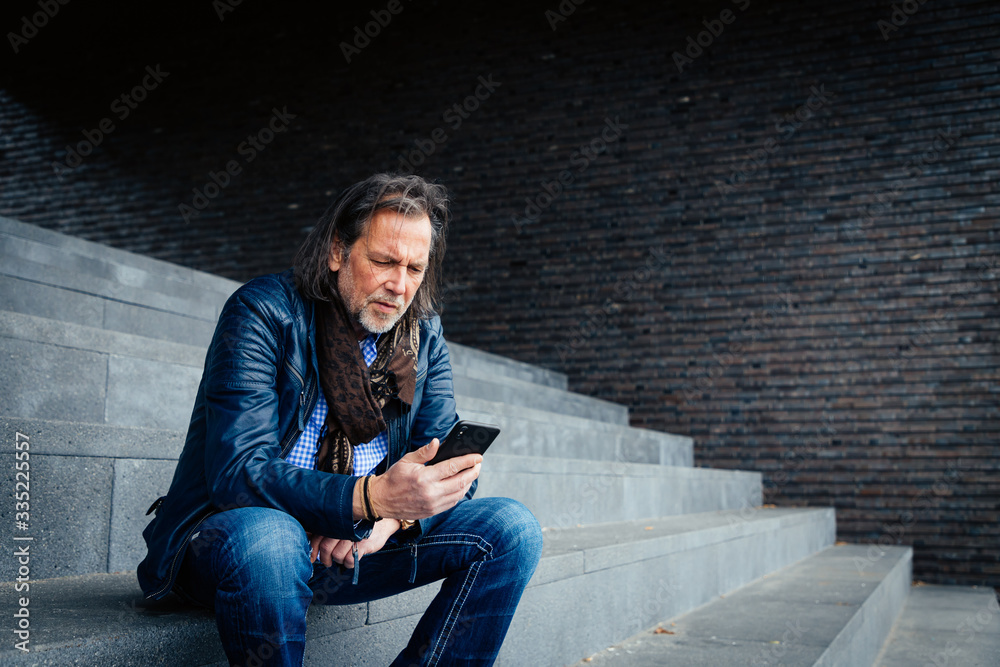 Older but cool man with beard and long grey hair uses a mobile phone in an urban environment