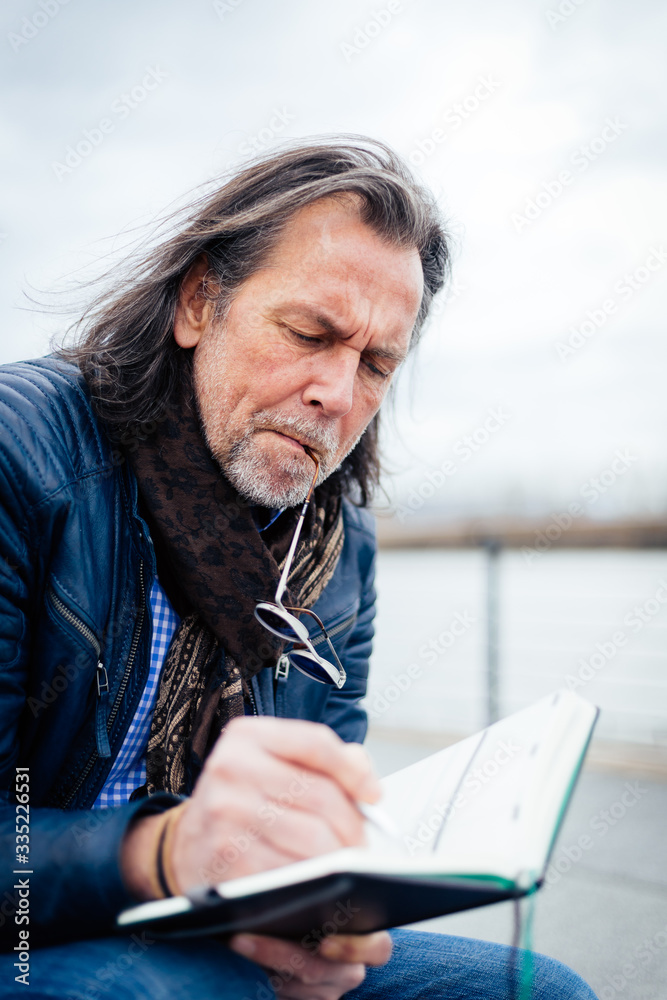 Older but cool man with beard and long grey hair writes in his notebook on the riverbank Older but cool man with beard and long grey hair writes in his notebook on the riverbank