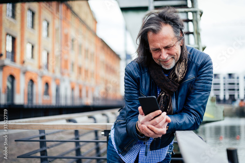 Older but cool man with beard and long grey hair uses a mobile phone in an urban environment
