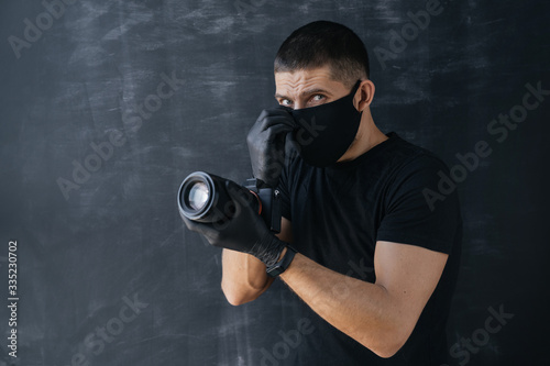 Portrait of a photographer, a man in a black medical mask and gloves, holding a professional digital camera and looking into the frame. Self-isolation and quarantine. Protection.
