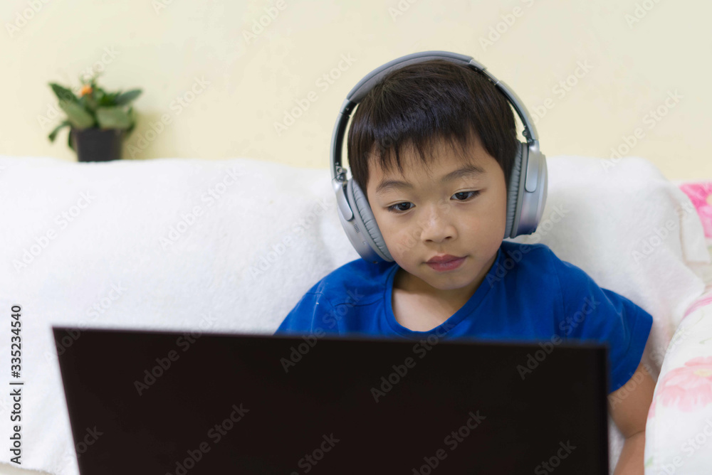 One boy is using digital screen computer with headphone to play game, do homework at home on sofa in dining room.Education school, technology, and computer learning during COVID-19 outbreak concept. One boy is using digital screen computer with headphone to play game, do homework at home on sofa in dining room.Education school, technology, and computer learning during COVID-19 outbreak concept.
