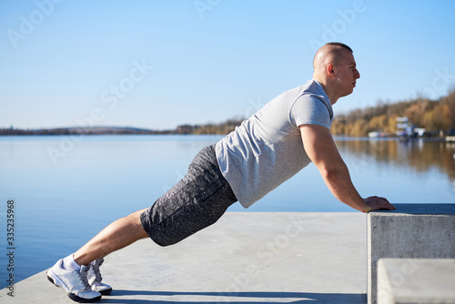Young sportsman, wearing grey outfit, is doing push-ups on concrete cube by the lake in the town.