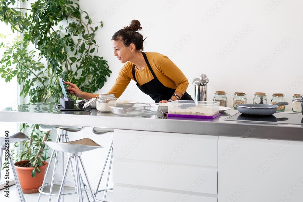 Young brunette woman looking at her digital tablet for a recipe on the web and cooking in a modern kitchen