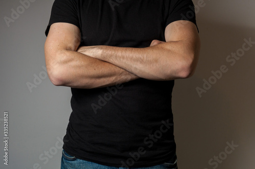 A man in a black T-shirt crossed his arms in front of his chest against a gray wall background