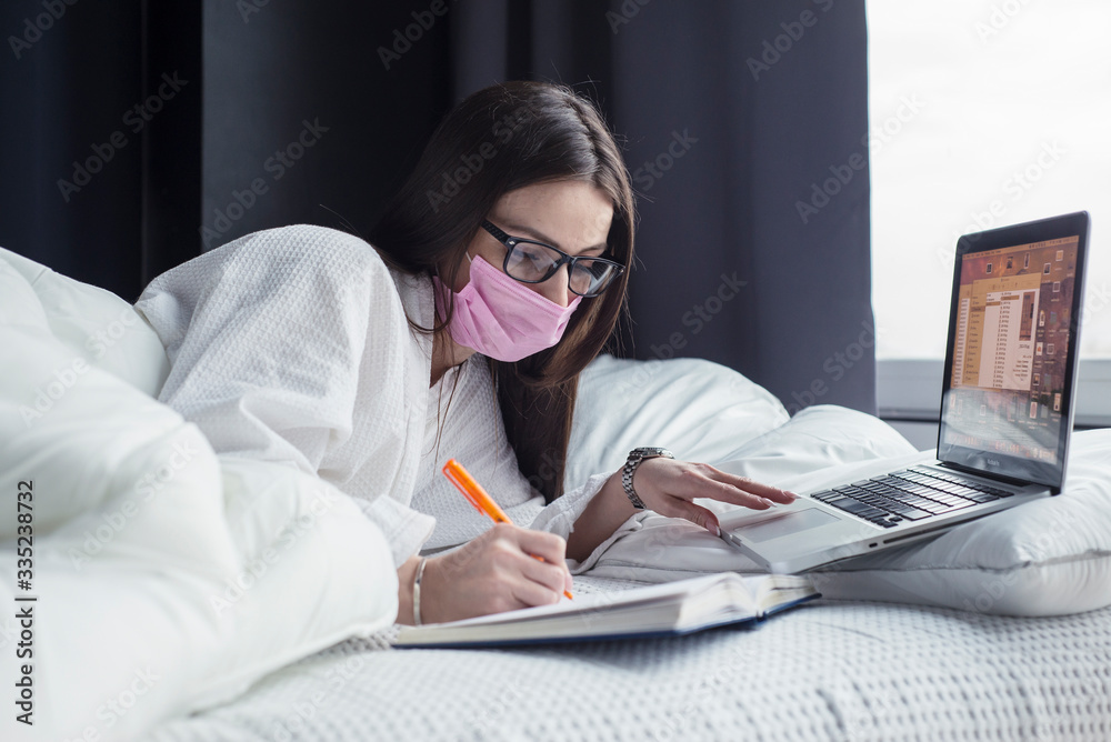 Obraz premium Quarantine, a woman in glasses and a mask lies in bed working on a laptop online. Stay at home.