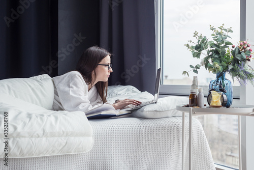 Quarantine, a woman with glasses lies in bed working on a laptop online. Stay at home.