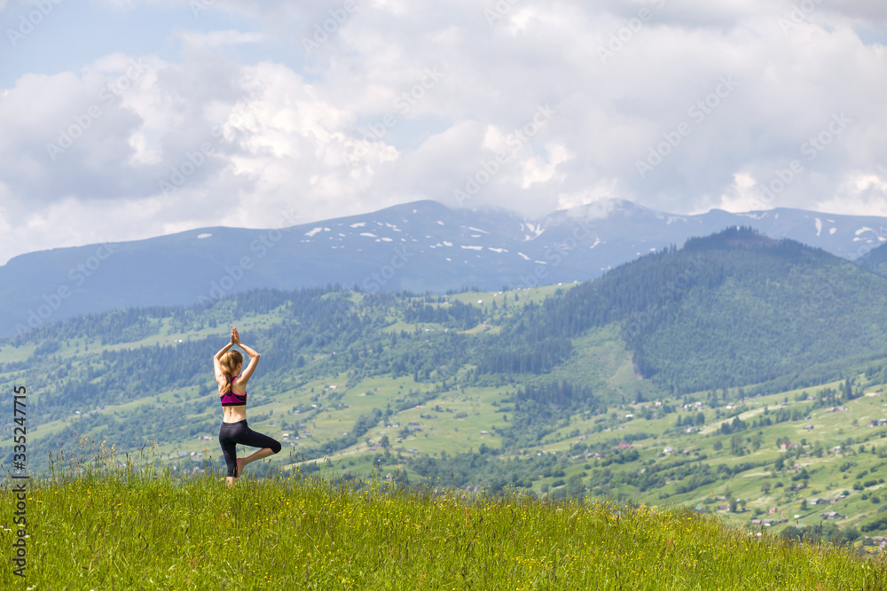 Attractive slim young woman doing yoga exercises outdoors on background of green mountains on sunny summer day.