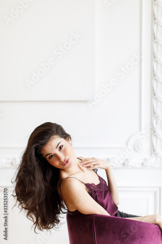 Brunette girl with thick long hair in a wine-colored blouse sits in an armchair against a white wall