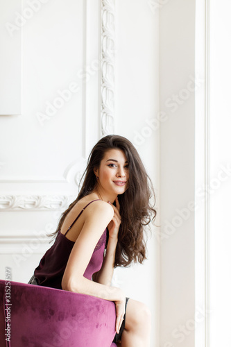 Brunette girl with thick long hair in a wine-colored blouse sits in an armchair against a white wall