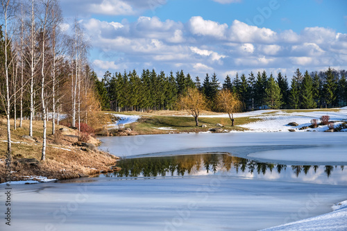 Spring landscape. Forest lake with reflection of clouds and trees growing along the shore, partially covered with ice and blue sky with clouds, at the beginning of a spring day.