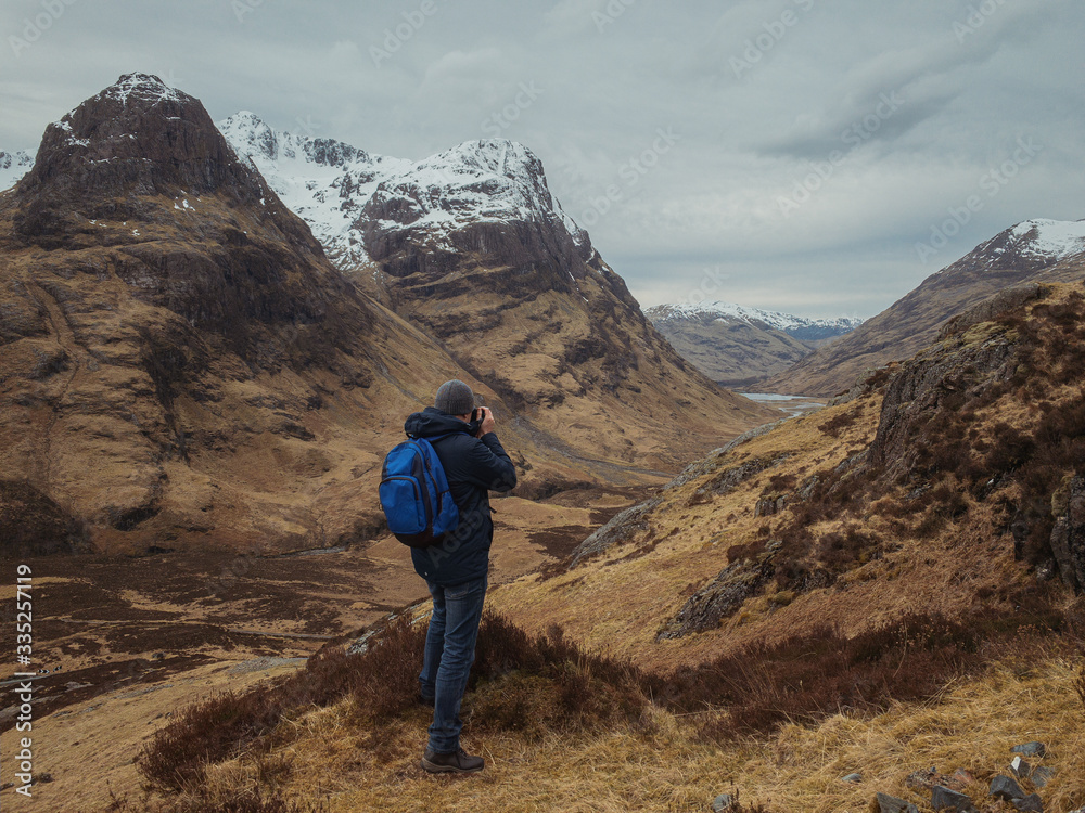 Man photographer with a backpack on the mountain from the back, looking at the mountain valley. The concept of active relaxation, motivation and goal achievement. Glencoe, Scotland, UK.