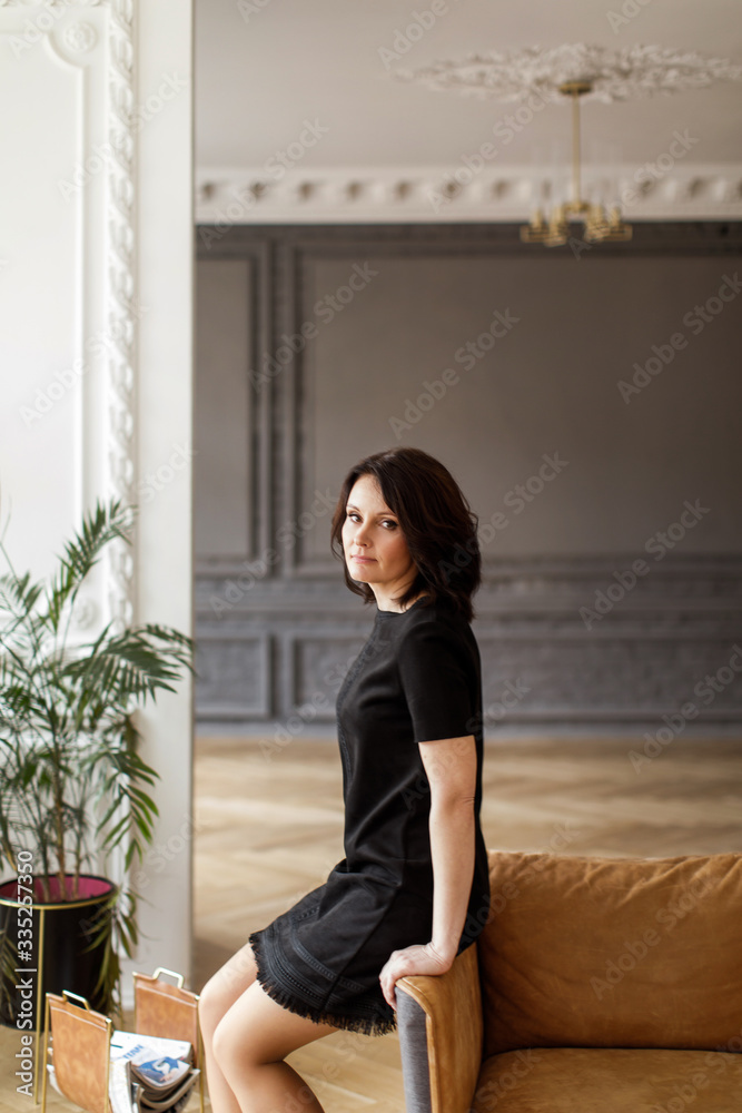Woman in a black dress sits on the back of a sofa in the interior Woman in a black dress sits on the back of a sofa in the interior