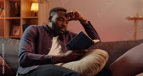 Handsome african guy leaning head with hand while sitting on sofa at living room. Young man spending free time while reading interesting book. Concept of leisure and hobby.