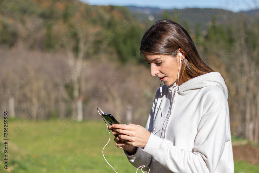 Surprised pretty young woman breathes fresh air in the park listening to music on headphones and holding a smartphone. She is wearing a light sweatshirt and has long brown hair. Surprised pretty young woman breathes fresh air in the park listening to music on headphones and holding a smartphone. She is wearing a light sweatshirt and has long brown hair.
