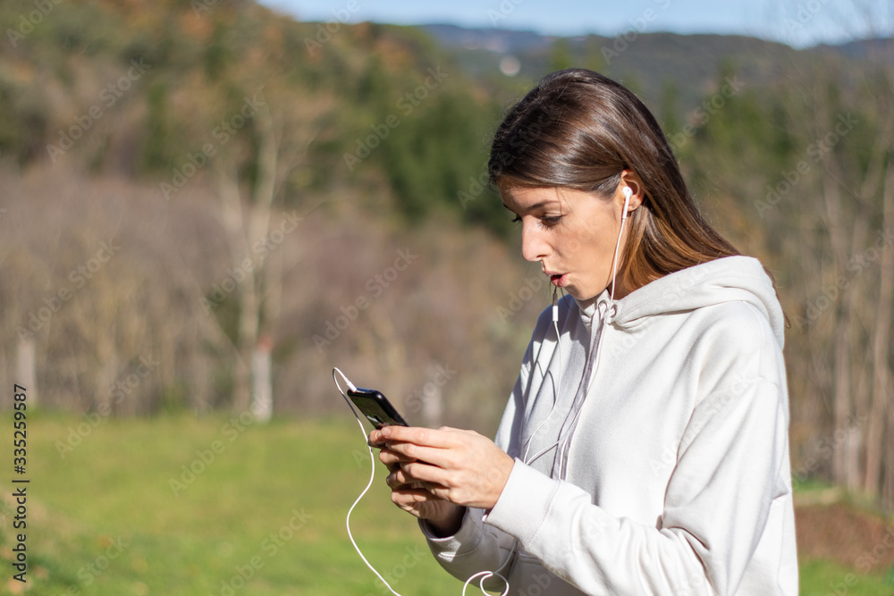 Shocked pretty young woman breathes fresh air in the park listening to music on headphones and holding a smartphone. She is wearing a light sweatshirt and has long brown hair.