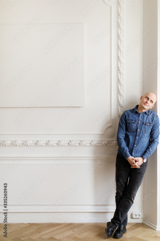 Portrait of a bald brutal Man in jeans standing against a white wall Portrait of a bald brutal Man in jeans standing against a white wall