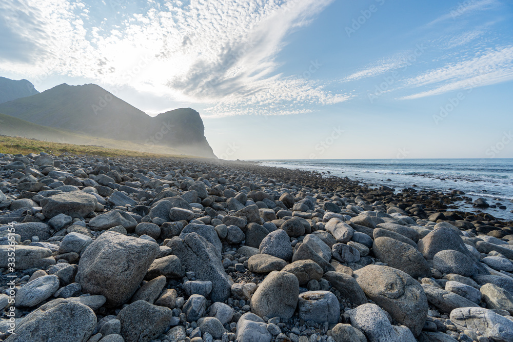 Pebble beach at Vestvågøy, Norway, with smooth stones leading to a calm sea and distant mountains under a sunny, cloud-streaked sky. Pebble beach at Vestvågøy, Norway, with smooth stones leading to a calm sea and distant mountains under a sunny, cloud-streaked sky.