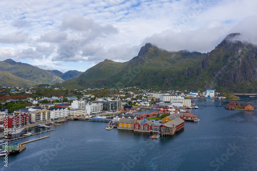 Coastal town in Vågan, Norway, with colorful houses lining the harbor, fishing boats and calm blue water set against rugged green mountains under a cloudy sky.