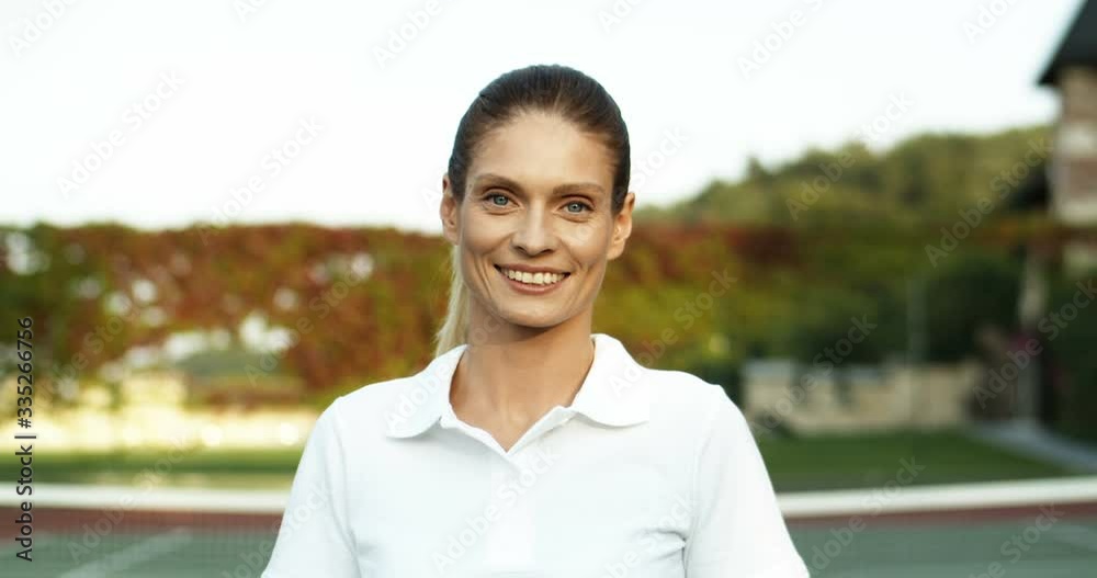 Close up of Caucasian beautiful young woman in white polo shirt smiling joyfully to camera on sunny day at green sport court. Portrait of pretty female tennis player outdoor. Close up of Caucasian beautiful young woman in white polo shirt smiling joyfully to camera on sunny day at green sport court. Portrait of pretty female tennis player outdoor.