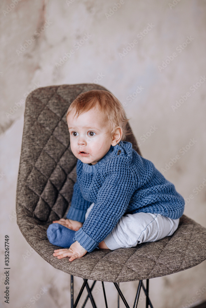 Cute Studio portrait of a one-year-old boy sitting on a chair. A boy in a blue sweater and beige pants Cute Studio portrait of a one-year-old boy sitting on a chair. A boy in a blue sweater and beige pants