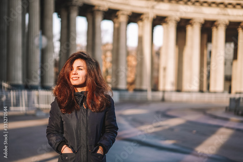 A young brunette girl with long hair in a jacket stands in the rays of the morning dawn on a deserted square in St. Petersburg against the background of the columns of the Kazan Cathedral