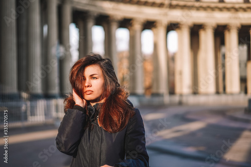 A young brunette girl with long hair in a jacket stands in the rays of the morning dawn on a deserted square in St. Petersburg against the background of the columns of the Kazan Cathedral