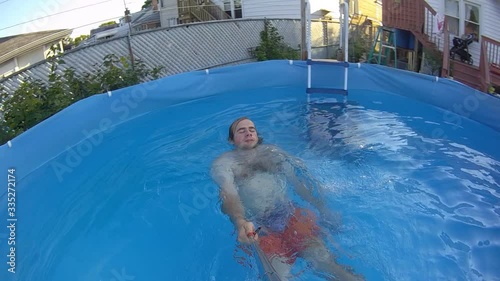 Slow motion of man in orange shorts recording hims self while swimming on his back in back yard swimming pool photo