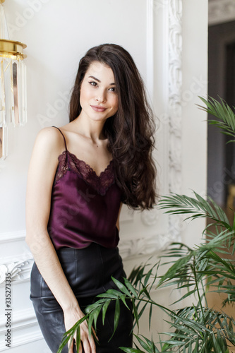 Joyful, beautiful girl laughs against the background of a white brick wall, portrait of an elegant brunette in the studio, the girl is hiding behind green plants, having fun, vertical photo © cmirnovalexander