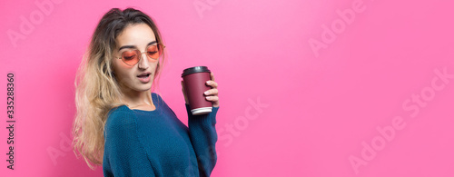 Glamor woman in glasses in a blue sweater with a drink of coffee on a pink background