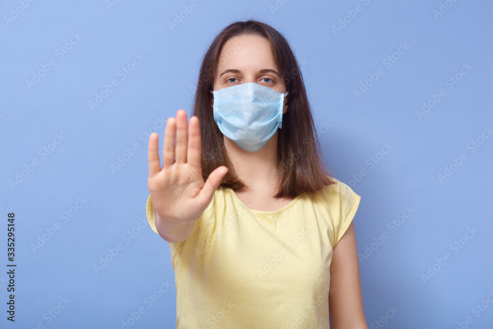 Indoor shot of Caucsian attractive woman wearing casual yellow t shirt and disposable medical mask, showing stop gesture with her palm, looking directly at camera. Covid 19, coronavirus concept. Indoor shot of Caucsian attractive woman wearing casual yellow t shirt and disposable medical mask, showing stop gesture with her palm, looking directly at camera. Covid 19, coronavirus concept.