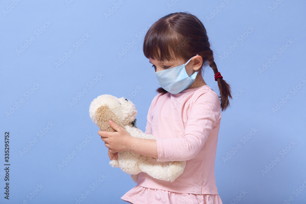 Close up portrait of little girl wearing casual clothing and medical mask, holding puppy toy in hands and looking at it, female child with pigtails isolated over blue studio background. Coronavirus. Close up portrait of little girl wearing casual clothing and medical mask, holding puppy toy in hands and looking at it, female child with pigtails isolated over blue studio background. Coronavirus.