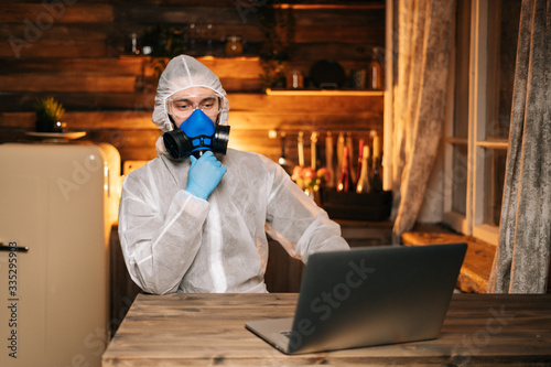 Young man wearing protective coveralls, goggles and medical respirator is typing on laptop. Concept of lifestyle in quarantine in Coronavirus COVID-19 Pandemic. photo