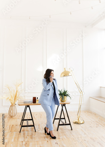 A serious business working freelance girl a woman with black hair in a pale blue suit working on a laptop and phone at a wooden Desk against a white wall in the home office