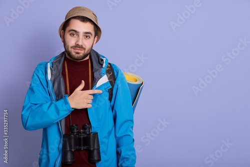 Close up portrait of bearded young traveller making gesture, pointing aside with forefinger, having binoculars around neck, wears cap and blue jacket. People, traveling and camping concept. Copy space