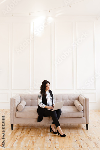 Serious business working freelance woman with black hair in a business suit working on a laptop and phone on a gray sofa against a white wall in the house, business portrait