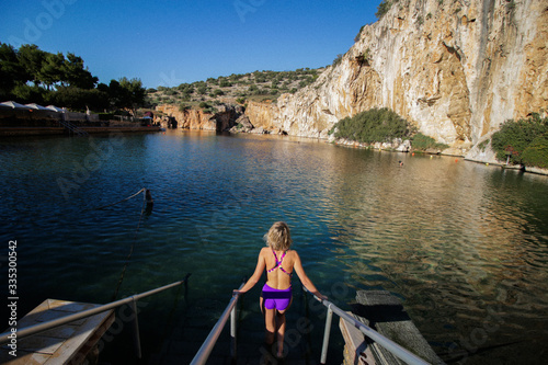 tanned girl in a swimsuit enters the lake against a background of stones and a photo taken from the back