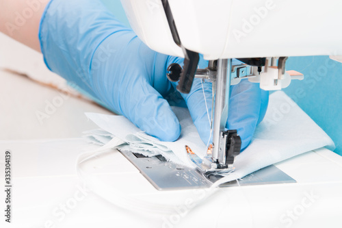 a woman in blue disposable medical gloves sews a protective mask for the face from cotton gray fabric on a sewing machine, blue background, the process of sewing masks, focus on fabric