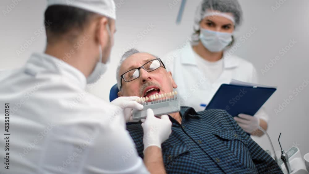 Male dentist choosing color of false jaw trying teeth to mature patient. Medium shot on RED camera