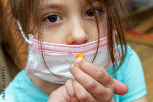 Child girl wearing medical protective mask with a pill in a hand at home during quarantine isolation photo