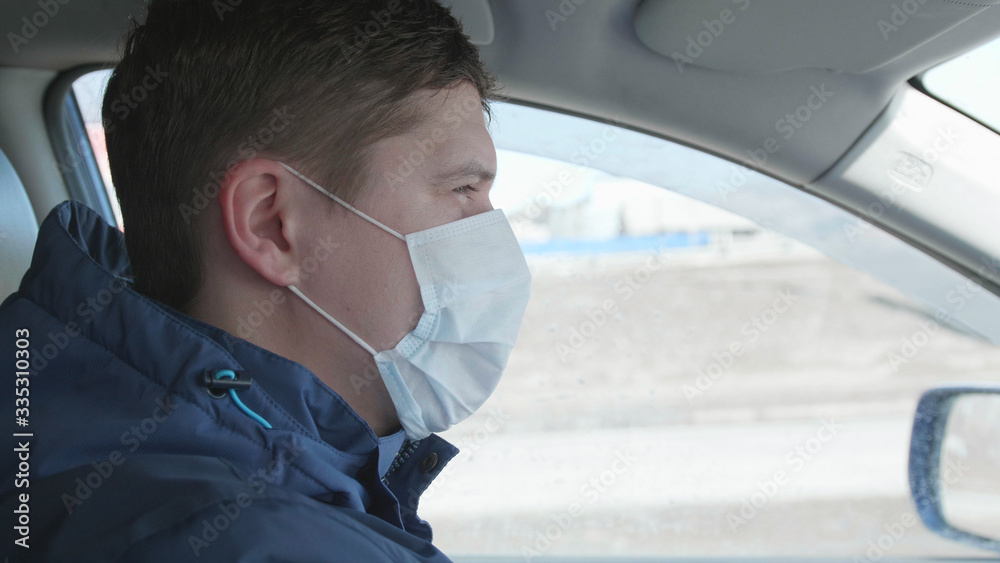 Guy is driving a car. The medical mask on the face. People use extra protection against coronavirus.