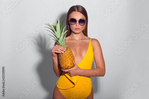 Sexy girl in yellow swimsuit and sunglasses posing with pineapple that covers her naked chest, isolated on white